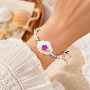 May include: A seashell bracelet featuring a large white shell with a purple flower accent. The bracelet includes colorful beads, pearls, and a small seashell charm. The bracelet is on a white cord.