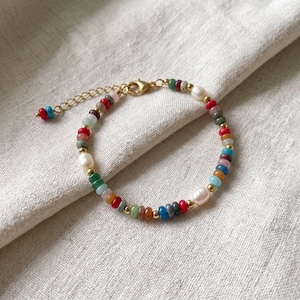 May include: A beaded bracelet featuring a repeating pattern of small, multi-colored round beads in shades of red, blue, green, and orange, interspersed with small gold-toned metal spacers and white pearls, finished with a gold-toned lobster clasp.