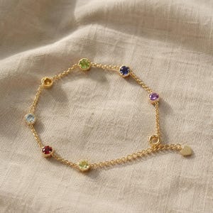 May include: A gold chain bracelet featuring seven round, multi-colored gemstones set in individual bezels. The stones include red, light blue, yellow, green, dark blue, purple, and pink hues. A small gold heart charm hangs near the circular clasp.