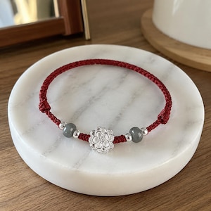 May include: A red cord bracelet featuring a central silver-toned flower charm, flanked by two smooth grey beads and four smaller silver-toned spherical beads. The bracelet is constructed with a braided red cord and adjustable knot closures.