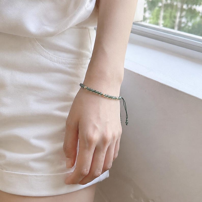 May include: A delicate green beaded bracelet with gold-colored accents. The bracelet is strung on a dark green cord and is adjustable. The bracelet is worn on a wrist, showcasing its minimalist design and natural color palette.