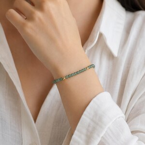 May include: A delicate bracelet featuring small, round, green beads strung together with gold-colored accents. The bracelet is worn on a wrist, showcasing its simple elegance and subtle color palette. The jewelry piece is a minimalist accessory.
