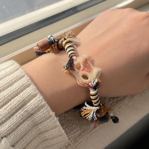 May include: A handmade bracelet featuring a cream-colored, bone-shaped bead with red markings. The bracelet has a woven cord with black, yellow, and white accents, and colorful tassel details. The bracelet is worn on a wrist.