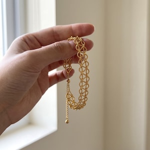 May include: A gold-toned bracelet featuring a delicate, interconnected lattice pattern made of small, textured beads. The bracelet includes an adjustable chain extension with a small spherical charm at the end, creating a flexible and intricate design.