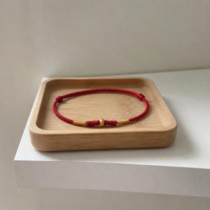 Può includere: Un bracciale in cordino rosso con una perlina centrale color oro affiancata da due piccole sezioni avvolte da filo dorato. Il bracciale è realizzato in sottile corda rossa intrecciata con nodi regolabili su entrambi i lati degli elementi dorati.