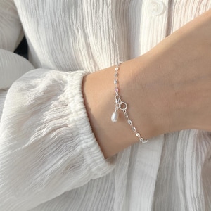 May include: A silver bracelet with a small pearl pendant. The bracelet has a chain of small, reflective links and a simple clasp. The pearl is white and teardrop-shaped. The bracelet is worn on a wrist.