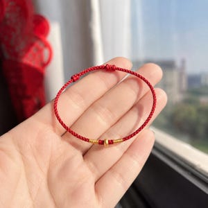 Puede incluir: Una pulsera de cord&oacute;n rojo fino con una peque&ntilde;a cuenta central de color dorado, flanqueada por dos breves envolturas de hilo met&aacute;lico dorado. La pulsera incluye dos nudos deslizantes ajustables en el cord&oacute;n para asegurar el ajuste.