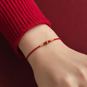 Puede incluir: Una pulsera de cord&oacute;n rojo fino con una peque&ntilde;a cuenta de color dorado en el centro, flanqueada por dos delicados envoltorios de hilo dorado. La pulsera est&aacute; hecha de un cord&oacute;n rojo trenzado y liso que rodea la mu&ntilde;eca con un dise&ntilde;o sencillo.