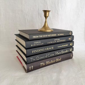 May include: A stack of five black hardcover books with gold lettering on the spines, topped with a small brass candlestick. The titles include From the Dust Returned, Jackal's Mistress, Finding Grace, The Queens of Crime, and The Blackest Bird.