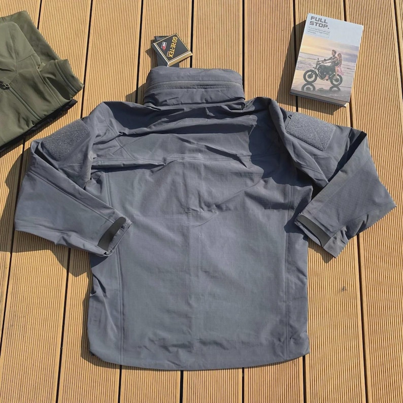 May include: A grey jacket with a high collar and multiple pockets. The jacket has a patch on the upper arm and black trim on the sleeves. A small book with the words "FULL STOP" is visible.