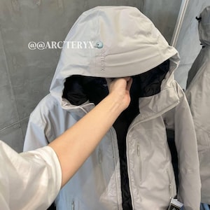 May include: A light gray hooded jacket with a black interior. The jacket has a zipper closure, multiple pockets, and the brand name "ARC'TERYX" printed on the upper left side. The jacket is displayed on a hanger.
