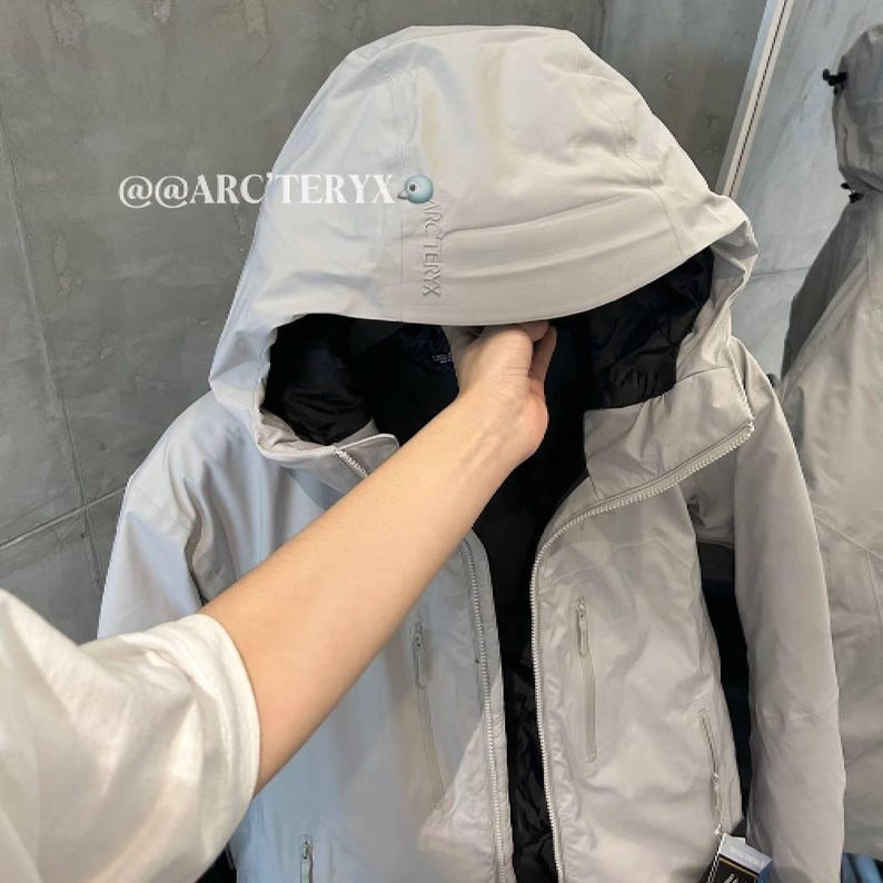 May include: A light gray hooded jacket with a black interior. The jacket has a zipper closure, multiple pockets, and the brand name "ARC'TERYX" printed on the upper left side. The jacket is displayed on a hanger.