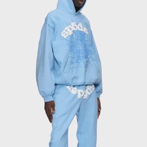 Op de afbeelding: Lichtblauwe hoodie en joggingbroek set. De hoodie heeft een witte spinnenwebafbeelding en het woord "sp5der". De joggingbroek heeft ook het woord "sp5der" erop gedrukt. De outfit is gemaakt van zacht materiaal.