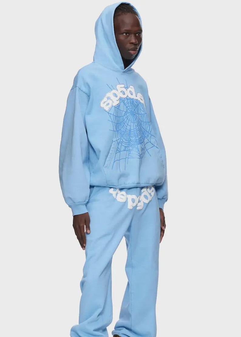 Op de afbeelding: Lichtblauwe hoodie en joggingbroek set. De hoodie heeft een witte spinnenwebafbeelding en het woord "sp5der". De joggingbroek heeft ook het woord "sp5der" erop gedrukt. De outfit is gemaakt van zacht materiaal.