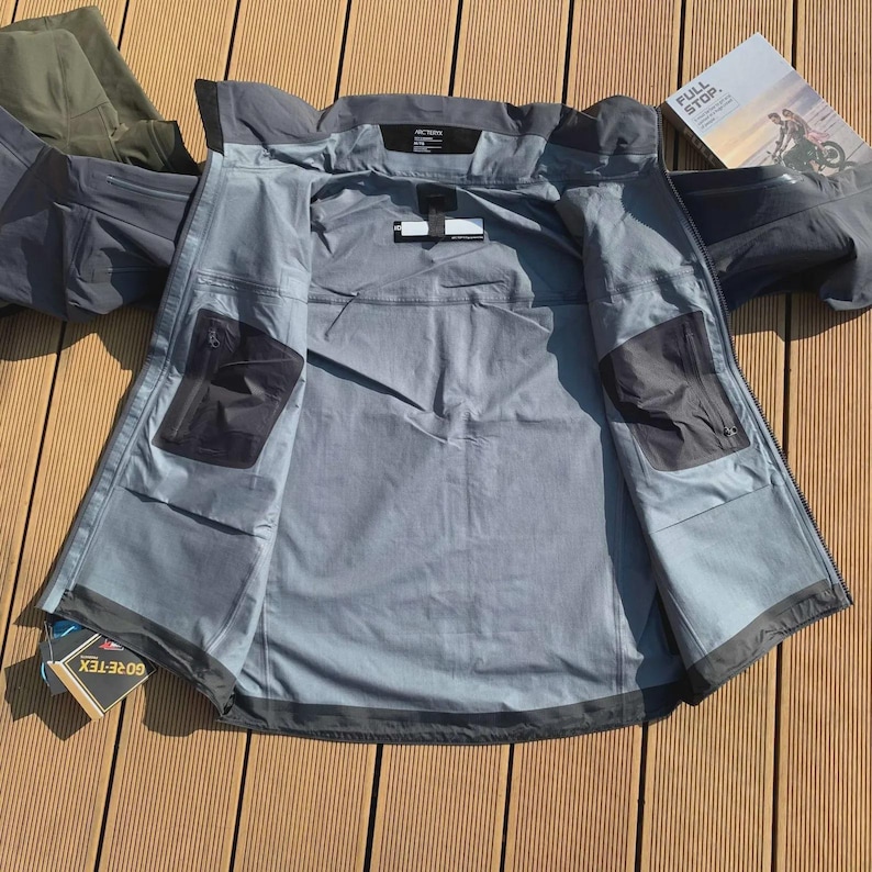 May include: A grey waterproof jacket, open to reveal the interior pockets and lining. The jacket is displayed on a wooden surface. A brochure with the text "FULL STOP" is visible, alongside a pair of green trousers.