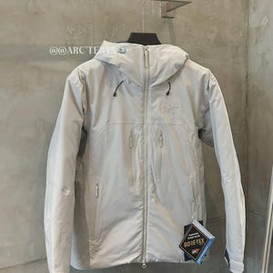 May include: A light gray hooded jacket with a full-length zipper and multiple front pockets. The jacket features the Arc'teryx logo and a Gore-Tex tag. The jacket is hanging on a rack against a neutral-colored wall.