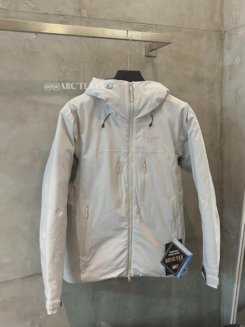 May include: A light gray hooded jacket with a full-length zipper and multiple front pockets. The jacket features the Arc'teryx logo and a Gore-Tex tag. The jacket is hanging on a rack against a neutral-colored wall.