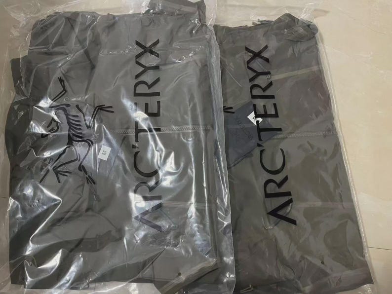 May include: Two grey jackets in clear plastic packaging. The jackets have the brand name "ARC'TERYX" printed vertically in large white letters. The brand's logo, a stylized bird fossil, is visible on one of the packages.