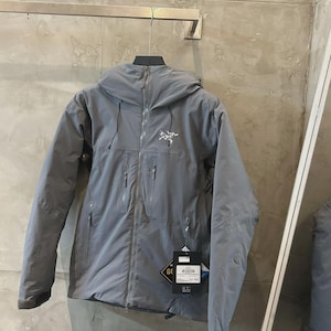 May include: A gray hooded jacket with a full-length zipper and multiple pockets. The jacket has a small logo on the chest and a tag with text. The jacket is hanging on a metal bar.