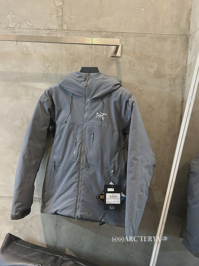 May include: A gray hooded jacket with a full-length zipper and multiple pockets. The jacket has a small logo on the chest and a tag with text. The jacket is hanging on a metal bar.
