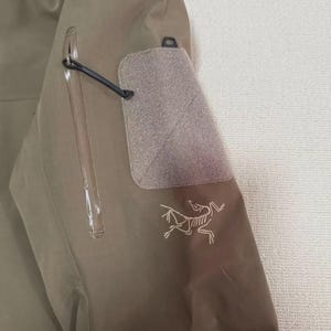 May include: Close-up of a tan outdoor jacket. Features include a zippered pocket, a velcro patch, and a small logo of a stylized animal. The jacket is made of a durable, water-resistant material.