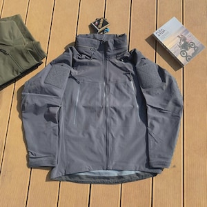 May include: A grey hooded jacket with a high collar and multiple zipped pockets. The jacket features a patch on the upper arm and a Gore-Tex tag. A book with the words "FULL STOP" is visible.