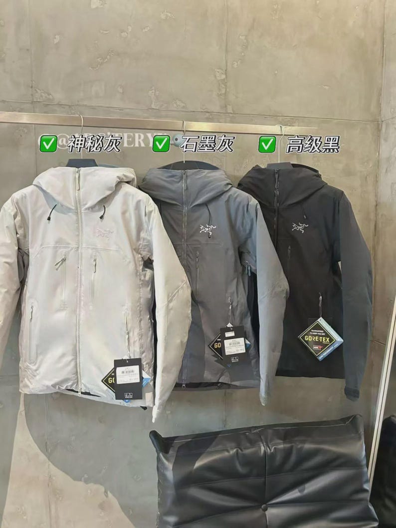 May include: Three hooded jackets in light grey, grey, and dark grey hang on a rack. Each jacket has a zipper closure and a small logo on the chest. The jackets have tags with the text "GORE-TEX". The text above the jackets reads "@Mysterious Grey ER", "Graphite Grey", and "High-end Black".