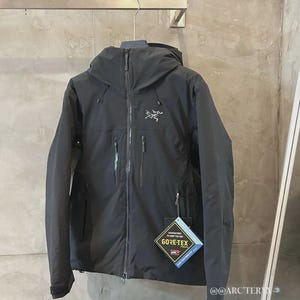 May include: Black hooded jacket with a zipper closure and multiple pockets. The jacket features the Arc'teryx logo on the chest and a Gore-Tex tag. The jacket is hanging on a metal bar.