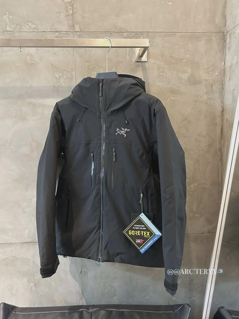 May include: Black hooded jacket with a zipper closure and multiple pockets. The jacket features the Arc'teryx logo on the chest and a Gore-Tex tag. The jacket is hanging on a metal bar.