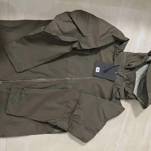 May include: An olive green hooded jacket with a full-length zip and a tag attached. The jacket features a drawstring hood and adjustable cuffs on the sleeves. The jacket is made of a lightweight material.