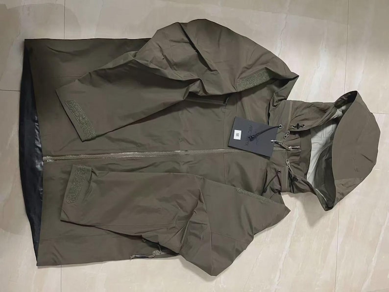 May include: An olive green hooded jacket with a full-length zip and a tag attached. The jacket features a drawstring hood and adjustable cuffs on the sleeves. The jacket is made of a lightweight material.