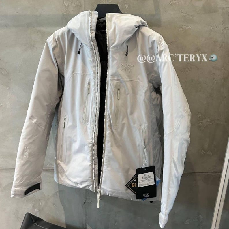 May include: A light gray hooded jacket with a black interior and multiple zippered pockets. The jacket has a tag attached with the text "GORE-TEX". The brand name "ARC'TERYX" is visible on the jacket.