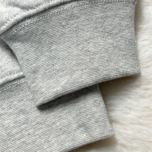Puede incluir: Primer plano del pu&ntilde;o de una sudadera gris claro. El pu&ntilde;o es acanalado y tiene una costura visible. La tela parece ser un material suave y tejido. El fondo es una superficie blanca texturizada y borrosa.