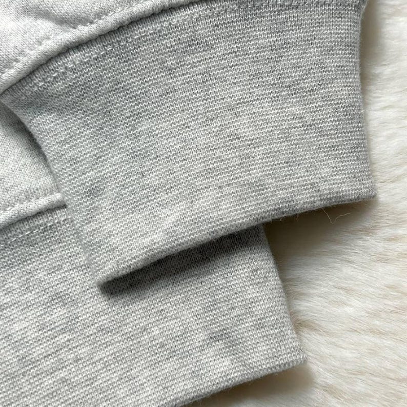 Puede incluir: Primer plano del pu&ntilde;o de una sudadera gris claro. El pu&ntilde;o es acanalado y tiene una costura visible. La tela parece ser un material suave y tejido. El fondo es una superficie blanca texturizada y borrosa.