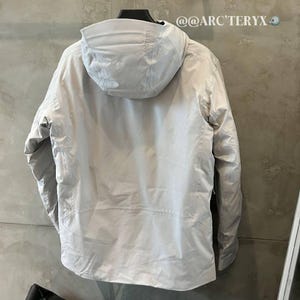 May include: A light grey hooded jacket with the text "ARC'TERYX" on the hood. The jacket is hanging on a black hanger against a grey wall. The jacket has a zipper and a pocket.