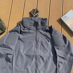 May include: A grey jacket with a high collar and multiple pockets. The jacket has a zip closure and a GORE-TEX tag. A book with the words "FULL STOP" is visible.