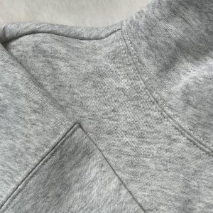 Puede incluir: Primer plano de una sudadera gris. La tela es un material suave y jaspeado. El cuello y las costuras son visibles, mostrando la construcci&oacute;n de la prenda. El aspecto general es informal y c&oacute;modo.