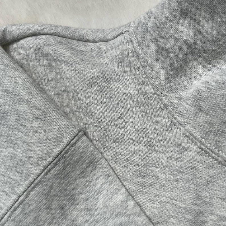 Puede incluir: Primer plano de una sudadera gris. La tela es un material suave y jaspeado. El cuello y las costuras son visibles, mostrando la construcci&oacute;n de la prenda. El aspecto general es informal y c&oacute;modo.