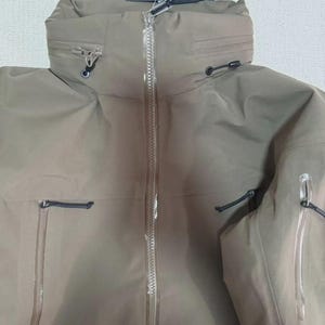 May include: A tan jacket with a high collar and a full-length zip. The jacket features multiple pockets and drawstrings. A tag with the brand name "Arc'teryx" is attached.