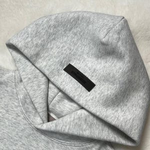 Puede incluir: Primer plano de una sudadera con capucha gris con capucha. Una etiqueta rectangular negra con la palabra "ESSENTIALS" est&aacute; cosida en la capucha. La sudadera con capucha est&aacute; hecha de una tela suave y texturizada.