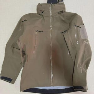 May include: A tan outdoor jacket with a high collar and multiple zipped pockets. The jacket has a waterproof design and features the brand name "Arc'teryx" on the sleeve. The jacket is designed for outdoor activities and protection from the elements.