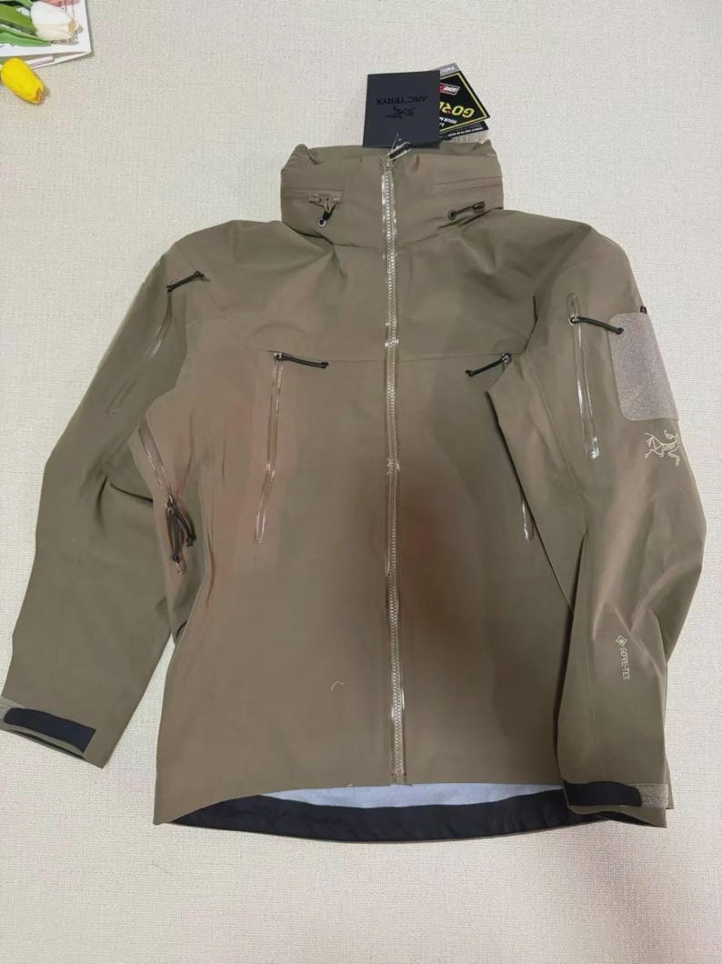 May include: A tan outdoor jacket with a high collar and multiple zipped pockets. The jacket has a waterproof design and features the brand name "Arc'teryx" on the sleeve. The jacket is designed for outdoor activities and protection from the elements.