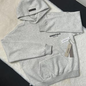 Puede incluir: Una sudadera con capucha gris claro con capucha y bolsillo delantero. La palabra "ESSENTIALS" est&aacute; impresa en el pecho. Una etiqueta que dice "PACSUN" est&aacute; adjunta a la sudadera con capucha.