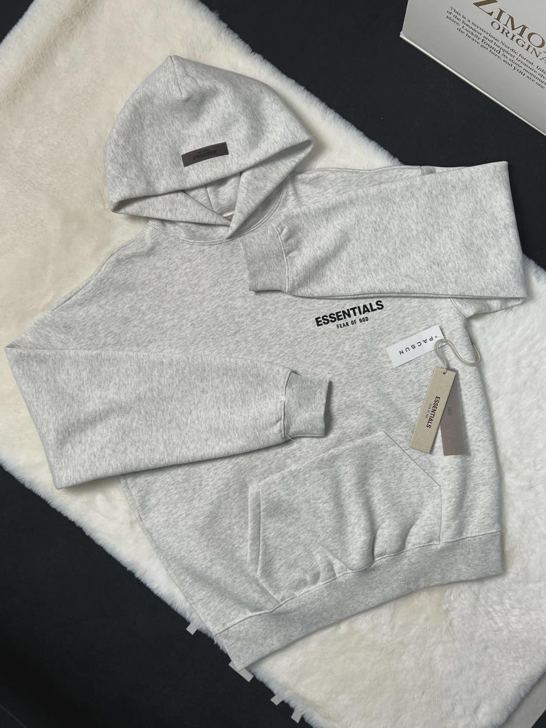 Puede incluir: Una sudadera con capucha gris claro con capucha y bolsillo delantero. La palabra "ESSENTIALS" est&aacute; impresa en el pecho. Una etiqueta que dice "PACSUN" est&aacute; adjunta a la sudadera con capucha.