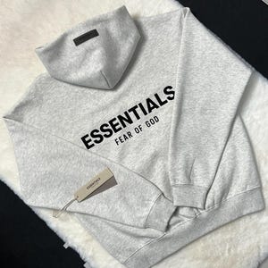Puede incluir: Una sudadera con capucha gris claro con la palabra "ESSENTIALS" impresa en negro en el pecho, con "FEAR OF GOD" debajo. La sudadera con capucha tiene una capucha a juego y una etiqueta que dice "ESSENTIALS".