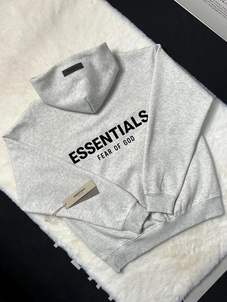 Puede incluir: Una sudadera con capucha gris claro con la palabra "ESSENTIALS" impresa en negro en el pecho, con "FEAR OF GOD" debajo. La sudadera con capucha tiene una capucha a juego y una etiqueta que dice "ESSENTIALS".