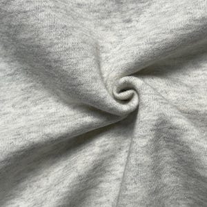 Puede incluir: Primer plano de una tela gris claro con un sutil patr&oacute;n moteado. La tela es suave y parece estar tejida, con textura y pliegues visibles. El material est&aacute; tejido apretado, creando una superficie lisa.
