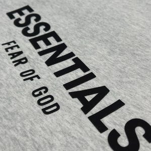 Puede incluir: Primer plano de una camiseta gris claro con la palabra "ESSENTIALS" impresa en letras negras grandes y en negrita. Debajo, el texto "FEAR OF GOD" est&aacute; impreso en una fuente m&aacute;s peque&ntilde;a.