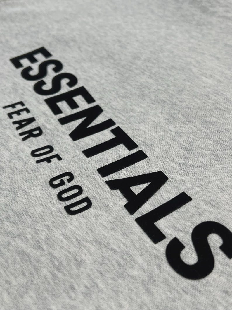 Puede incluir: Primer plano de una camiseta gris claro con la palabra "ESSENTIALS" impresa en letras negras grandes y en negrita. Debajo, el texto "FEAR OF GOD" est&aacute; impreso en una fuente m&aacute;s peque&ntilde;a.