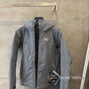 May include: A gray hooded jacket with a zipper closure and multiple pockets. The jacket has a white logo on the chest and a Gore-Tex tag. The jacket is hanging on a metal bar.